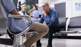 A Stressed Senior Man Sitting in a Waiting Room With Two Physicians Talking To Each Other in the Background What Do You Do if Your Doctor Isn’t Helping You