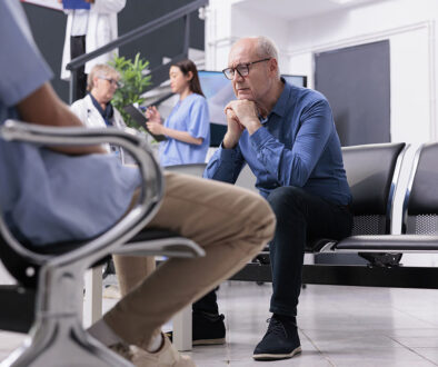 A Stressed Senior Man Sitting in a Waiting Room With Two Physicians Talking To Each Other in the Background What Do You Do if Your Doctor Isn’t Helping You