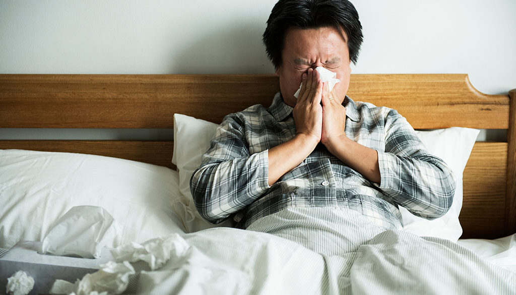 An Asian Man Sitting in Bed Blowing His Nose Hay Fever vs Cold Symptoms