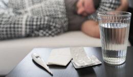 Closeup Of A Nightstand With A Glass Of Water, Medicine, & Thermometer On It & A Man Laying In Bed In The Background Prevent Flu Dehydration